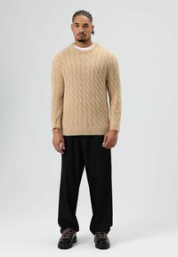 Beige gebreide trui met kabelpatroon, ronde hals en lange mouwen. Gecombineerd met losse zwarte broek en zwarte schoenen met roze veters.