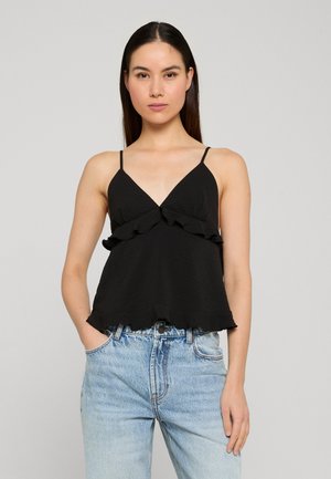 Kvinde med langt mørkt hår iført sort ærmeløs top med flæser og lyseblå, højtaljede jeans, stående med den ene hånd i lommen.