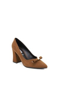 Scarpa da donna in suede marrone con tacco alto e punta affusolata, dotata di un dettaglio a fiocco e perle sulla tomaia. Tacco robusto per maggiore stabilità.