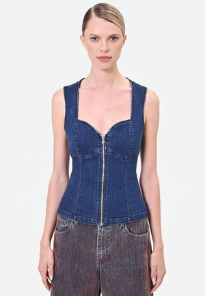 Femme portant un top en denim bleu foncé ajusté sans manches avec une fermeture éclair devant, associé à un pantalon en denim large et délavé.