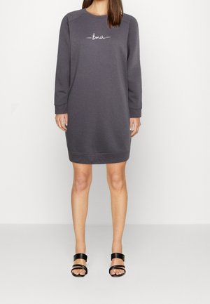 Kobieta w ciemnoszarej sukience typu sweatshirt do kolan z długim rękawem, z napisem "Bench" na piersi, oraz w czarnych sandałach z odsłoniętymi palcami.