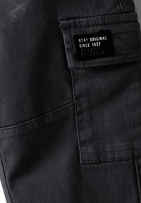 Svart denimtyg med en texturerad yta, med en rektangulär patch med text. Inkluderar synliga sömmar och en sidoficka.