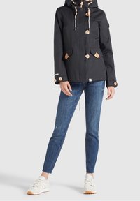 Veste imperméable noire avec boutons en cuir marron clair, capuche à cordon de serrage et deux poches avant, associée à un jean skinny bleu et des baskets blanches.