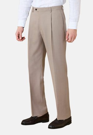 Mann trägt beigefarbene plissierte Anzughose, weißes, gestreiftes Langarmhemd und dunkelbraune Lederloafer, steht vor weißem Hintergrund.