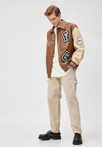 Bruin leren varsityjack met crèmekleurige mouwen, voorzien van grote geborduurde letters en graphics. Gemaakt in combinatie met lichtbeige broek en zwarte schoenen.