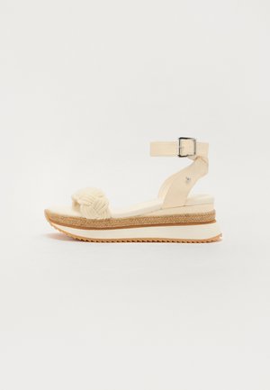 Cream wedge sandal with a woven knot detail on the strap, ankle buckle, and textured jute midsole. Non-slip rubber outsole.