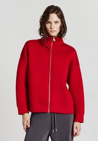 Felpa rossa con zip e colletto alto, dalla forma leggermente ampia, spalle basse e polsini a costine. Abbinata a pantaloni grigi con cordino.