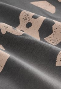 Tessuto grigio con lettering rosa sbiadito e strutturato, insieme a forme astratte. Il materiale sembra morbido e leggermente stropicciato, mostrando un design informale.