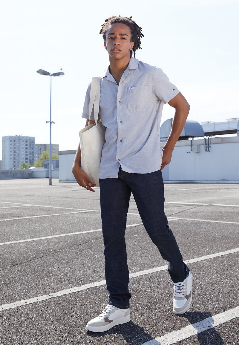 Lichtgrijs overhemd met korte mouwen en knopen, donkerblauwe jeans en witte sneakers met grijze accenten. Model houdt een beige draagtas vast. Buitensetting.