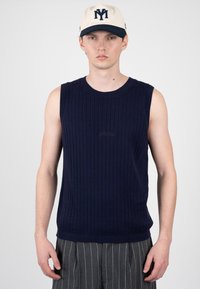 Jeune homme portant une casquette de baseball blanche et bleu marine, un pull sans manches côtelé bleu marine et un pantalon gris à fines rayures, debout devant un fond uni.