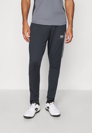 Mężczyzna w czarnych spodniach dresowych Under Armour, szarej koszulce i białych butach sportowych stoi na szarej podłodze na tle białej ściany.
