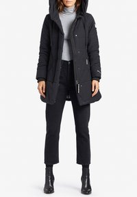 Svart vadderad parkas med huva, med en quiltad design, frontdragkedja och sidofickor, tillsammans med en grå polotröja och svarta jeans.