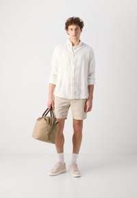 Chemise en lin blanche légère, shorts texturés beige, baskets beige, portant un sac en simili cuir tan avec des poignées sombres. Palette de couleurs simple et neutre.