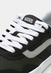 Mörkgrön mockasneaker med vita snörning, en grå sidrand och en svart Vans-logga på tungan. Texturerad ovandel med perforeringar.
