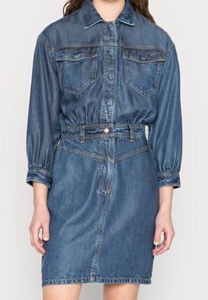 Kvinde iført en blå denimkjole med lange ærmer, knaplukning foran, brystlommer og en figursyet talje med bæltestropper.