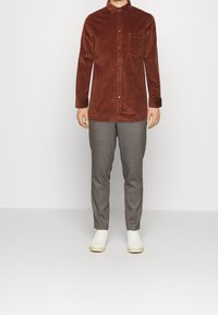 Hnědá manšestrová košile s designem na knoflíky, jednou náprsní kapsou, v kombinaci se šedými kalhotami a bílými teniskami. Jednoduché pozadí.