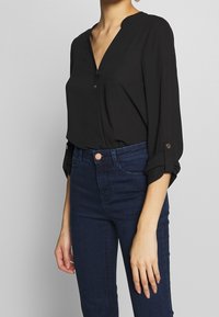 Blouse noire avec un décolleté en V et des détails de boutons, manches longues retroussées aux poignets. Associée à un jean fitted bleu foncé, mettant en valeur une texture lisse.