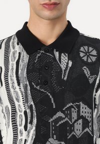 Svart-hvitt mønstret polo skjorte med tre knappåpninger, teksturert stoff og solid svart krage med geometriske design.