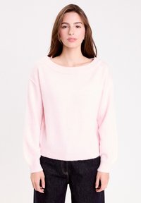 Pull en maille rose clair avec un col large et des manches ballon, présentant une texture douce et des bords côtelés. Associé à un jean foncé.