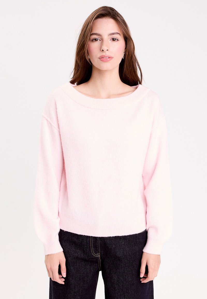 Pull en maille rose clair avec un col large et des manches ballon, présentant une texture douce et des bords côtelés. Associé à un jean foncé.