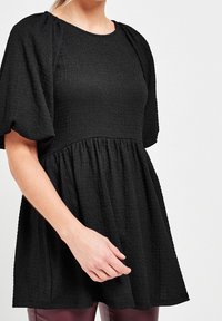 Haut noir texturé avec des manches bouffantes courtes et une taille froncée, présentant un col rond et une silhouette fluide.