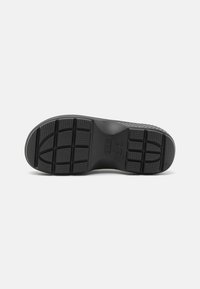 Semelle de chaussure en caoutchouc noir avec un motif texturé, présentant des blocs de bande de roulement proéminents et un bord lisse. Comprend des éléments circulaires au centre.