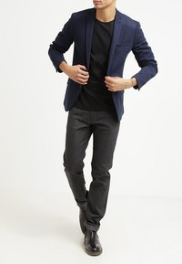 Mörkblå kavaj med strukturerade axlar, enkel bröstficka, bärs över en svart skjorta, matchad med mörkgrå jeans och svarta stövlar.