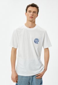 Hvid bomulds t-shirt med korte ærmer, der har et farverigt spiral grafisk design på venstre brystområde. Casual pasform, rund halsudskæring.