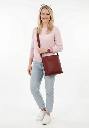 Lächelnde Frau mit Brille auf dem Kopf, rosa Pullover, helle Jeans, weiße Sneakers, lila Uhr, trägt eine große braune Leder-Umhängetasche.