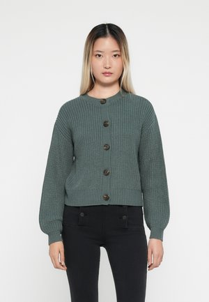 Grønn strikket cardigan med rund hals og syv store knapper. Har lange pufferm og ribbestrikket tekstur i nederkant og på mansjettene.