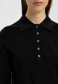 Polo shirt nero in maglia con colletto a punta, placket con sei bottoni e bottoni grigio chiaro. Texture liscia con un design aderente.