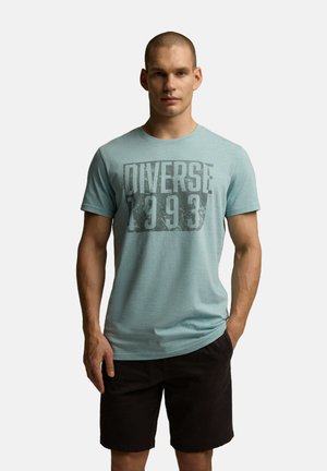 Jasnoniebieska koszulka z krótkim rękawem z teksturowanym pasowym wzorem, z dużym szarym nadrukiem na środku mówiącym "DIVERSE 1993".