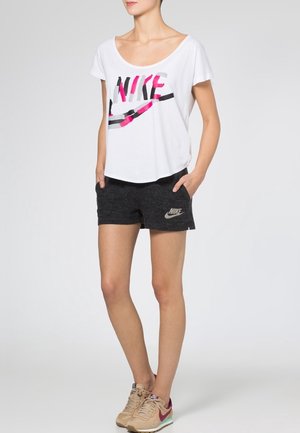 Bílý tričko Nike s barevným grafickým potiskem, spojené s černými šortkami s malým logem. Model má na sobě béžové a vínové sportovní boty.