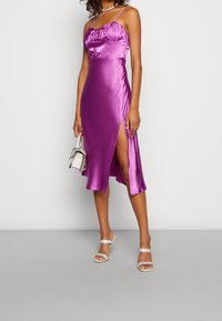 Robe en satin violet avec bretelles spaghetti réglables, corsage froncé et fente latérale. Associée à des talons argentés à lanières et un petit sac à main.