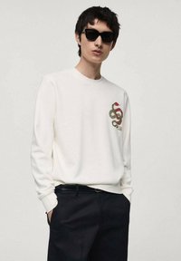 Witte sweatshirt met lange mouwen, voorzien van een groene en rode slangafbeelding op de borst, gecombineerd met donkere broeken en een zwarte zonnebril.