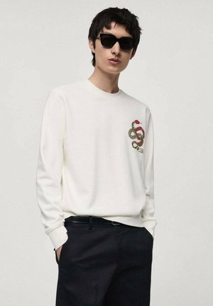 Weißes Sweatshirt mit langen Ärmeln und einem grünen und roten Schlangenmotiv auf der Brust, kombiniert mit dunklen Hosen und schwarzen Sonnenbrillen.