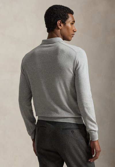 Polo grigia a maniche lunghe con colletto, con orlo a coste. Abbinata a pantaloni grigio scuro, che mostrano un tessuto strutturato.