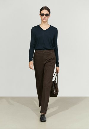 Donna con capelli corti e scuri che indossa occhiali da sole, una maglia nera a maniche lunghe, pantaloni marroni, scarpe nere e porta una borsa marrone.