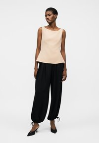 Žena stoji noseći bezrukavu bež majicu, široke crne hlače skupljene na gležnjevima i crne cipele s špicastim vrhovima protiv jednostavne bijele pozadine.