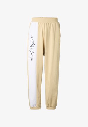 Beežid dressipüksid, millel on elastne vöökoht ja mansetid ning vasakul jalal valge vertikaalne triip, millel on stiliseeritud must tekst.