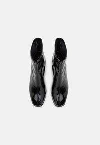 Paire de bottines noires brillantes avec bouts pointus et talons courts, vues de dessus sur un fond blanc uni.