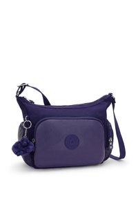 Fialová nylonová crossbody taška s texturovaným povrchem, přední zipová kapsa a malý chlupatý přívěsek ve tvaru opice. Součástí je nastavitelné rameno.