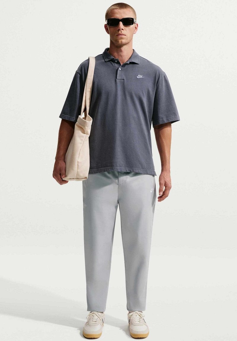 Uomo con occhiali da sole neri, polo Nike grigio scuro, pantaloni Nike grigio chiaro, sneakers bianche, con borsa beige a tracolla.
