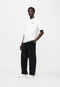 Λευκό κοντομάνικο t-shirt με μαύρο λογότυπο, συνδυασμένο με μαύρο παντελόνι που έχει κόκκινες λεπτομέρειες και λογότυπο. Συμπληρώνεται με λευκά αθλητικά παπούτσια.