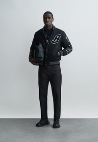 Muž oblečený v čiernej varsity bunde s hviezdičkovými nášivkami, čiernych džínsoch a čiernych teniskách, drží čierny batoh na jednoliatom pozadí.