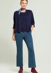Cardigan navy con bottoni, abbinato a un top navy, jeans a zampa blu e scarpe bordeaux, accentuato da una sciarpa blu con motivo.
