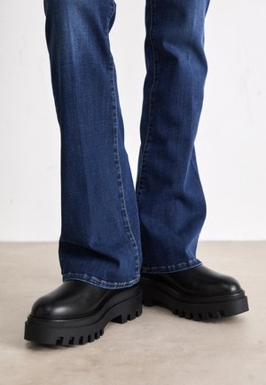 Stivali platform neri chunky con suole in gomma testurizzata, punta arrotondata e tomaia in pelle liscia, abbinati a jeans svasati blu.