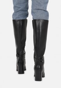 Bottes noires en cuir montant jusqu'au genou avec un talon carré, des accents de fermeture éclair et un détail de sangle à boucle, vues de dos avec un pantalon en jean.