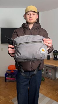 Grijze stoffen schoudertas met een ritssluiting aan de bovenkant, verstelbare band en een klein oranje logo. De tas heeft een gladde textuur en een rechthoekige vorm.