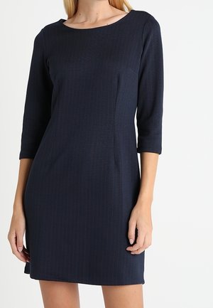 Granatowa sukienka długości do kolan z delikatnymi prążkami, rękawami do łokcia i okrągłym dekoltem, noszona przez stojącą kobietę.
